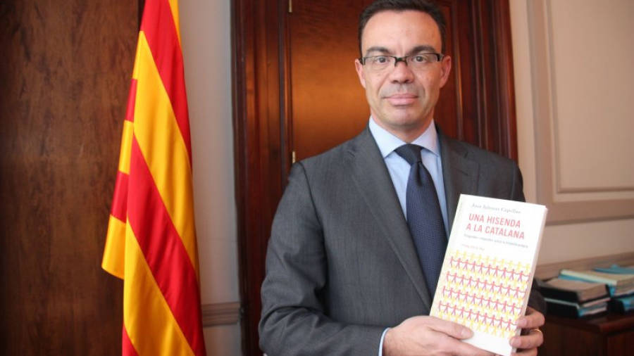 L'assessor del president de la Generalitat Joan Iglesias, amb el seu nou llibre a les mans, 'Una hisenda a la catalana' i la bandera catalana del seu despatx al fons. Foto: ACN