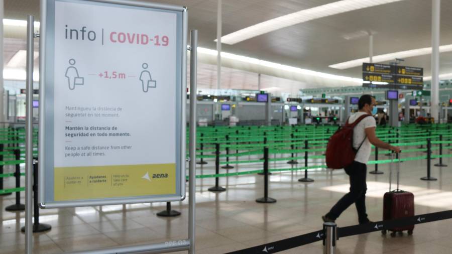 Pla general de un cartel informativo de la covid-19 y de un pasajero arrastrando una maleta con el control de seguridad de la T1 de l'Aeroport del Prat. Foto: ACN